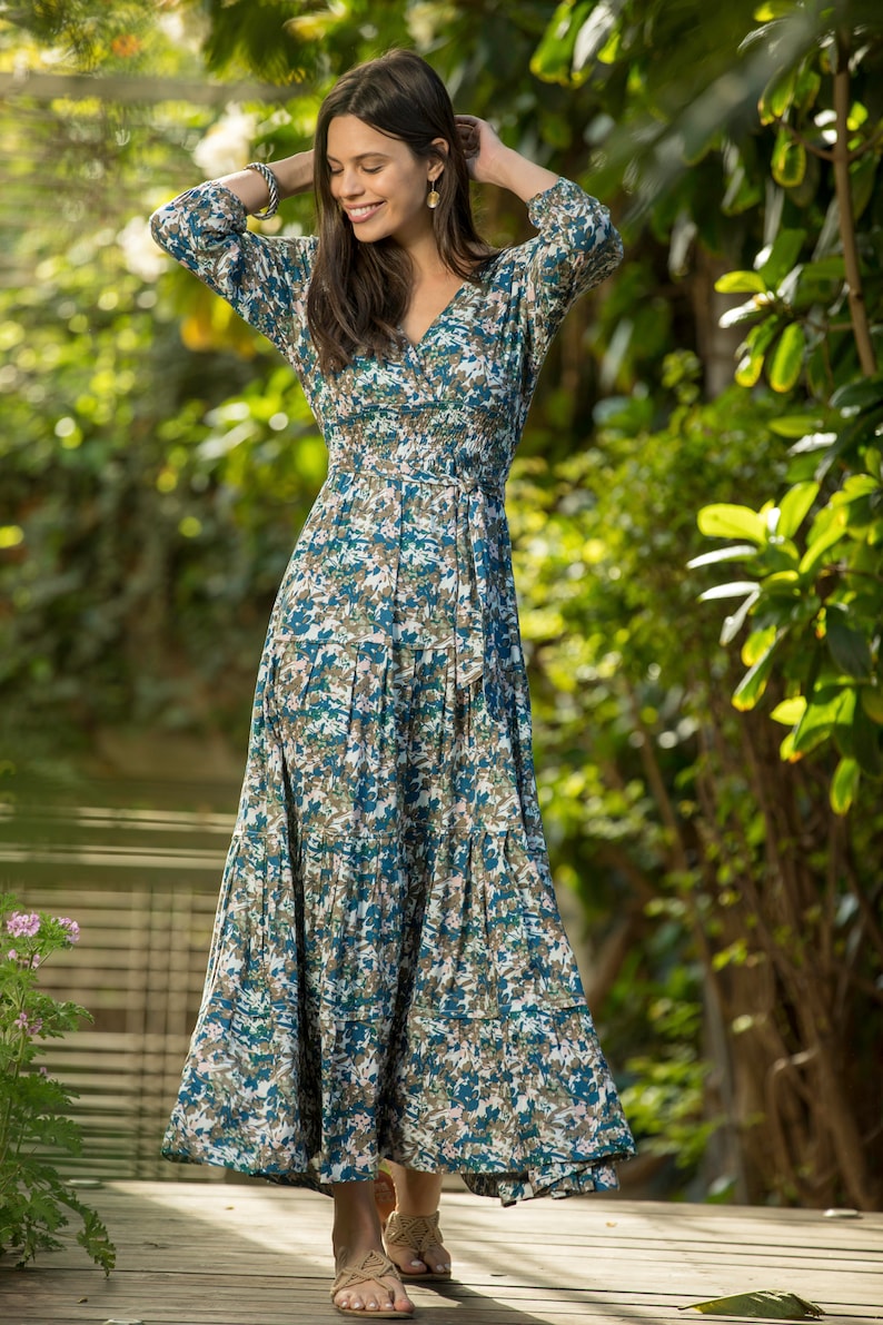 May include: A woman wearing a long, flowing blue and white floral dress with a cinched waist and a tie belt. She is standing on a wooden deck with green foliage in the background.