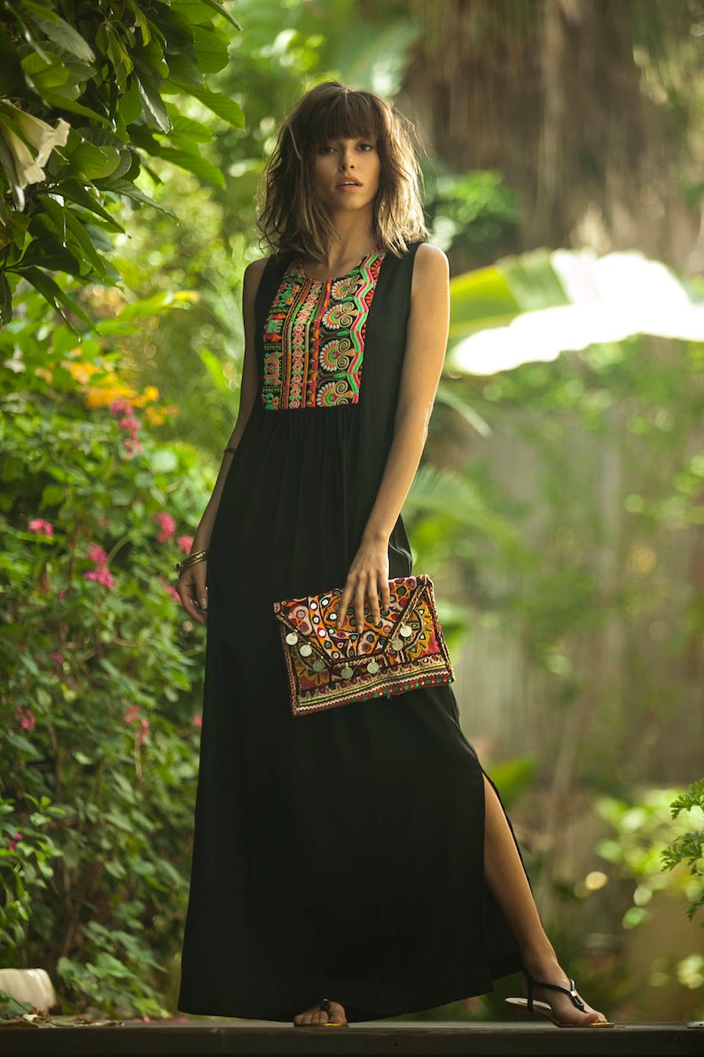 May include: A woman wearing a black maxi dress with colorful embroidery on the bodice. She is holding a colorful clutch bag with mirror accents.