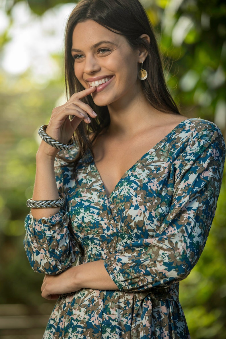 May include: A woman wearing a blue and brown floral print dress with a v-neckline and long sleeves. She is wearing two silver bracelets on her left wrist and a pair of gold earrings.