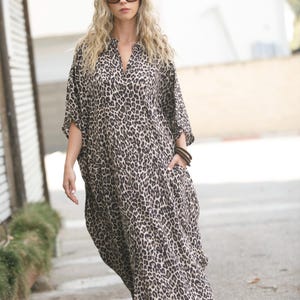 May include: A loose-fitting, floor-length dress featuring a brown and black leopard print pattern. The garment has a V-neckline, three-quarter length sleeves, and two side pockets. The fabric drapes loosely over the body, creating a relaxed silhouette.