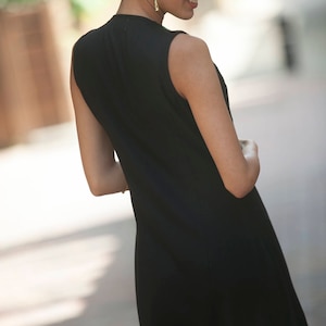 May include: A woman wearing a black sleeveless dress and sunglasses. She is standing on a sidewalk and looking down.