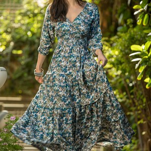 May include: A woman wearing a long, flowing blue and green floral dress with a cinched waist and a tiered skirt. She is standing on a wooden deck with greenery in the background.