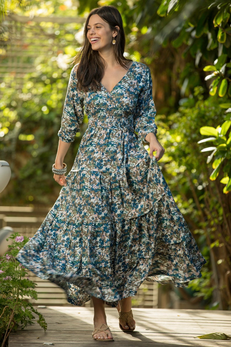 May include: A woman wearing a long, flowing blue and green floral dress with a cinched waist and a tiered skirt. She is standing on a wooden deck with greenery in the background.