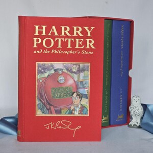 May include: A red hardback book titled "Harry Potter and the Philosopher's Stone" with gold lettering, featuring an illustration of the Hogwarts Express train. Two other books are visible in a box set. A small owl figurine is also present.