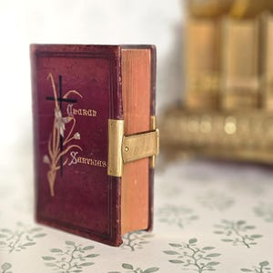 May include: A maroon book with gold lettering and a gold clasp. The cover features a cross and floral design with the words "Oraran Services". The book's pages have a golden hue.
