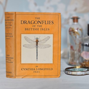 May include: An antique book titled "The Dragonflies of the British Isles" by Cynthia Longfield. The cover is orange with black text and a detailed illustration of a dragonfly. The book is displayed with a magnifying glass and glass bottles in the background.