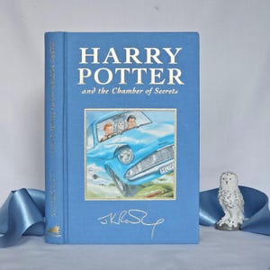 May include: A light blue hardcover book titled "Harry Potter and the Chamber of Secrets" with gold lettering. The cover shows a blue car with two people inside. A small white owl figurine is positioned to the right of the book.