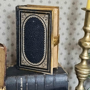 May include: A miniature antique book with a dark blue leather cover and gold detailing. The cover has an oval design with intricate patterns. The book is open, showing aged, yellowed pages. A gold clasp secures the book. The book is placed on top of another book.