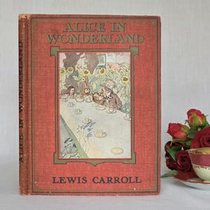 May include: A vintage hardcover book titled Alice in Wonderland by Lewis Carroll. The red cover features a central illustration of a tea party scene with the Mad Hatter and other characters, framed by green borders and dark red textured fabric.