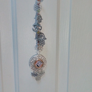 May include: A silver-toned spiritual wall hanging with various charms, including a Hamsa hand, the Om symbol, and a lotus flower. The hanging also features pink and green beads, and a decorative butterfly charm.