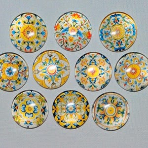 May include: A collection of ten round decorative glass cabochons. Each cabochon features a unique, colorful floral design with yellow, blue, and orange hues on a white background. The designs are intricate and detailed, resembling traditional tile patterns.