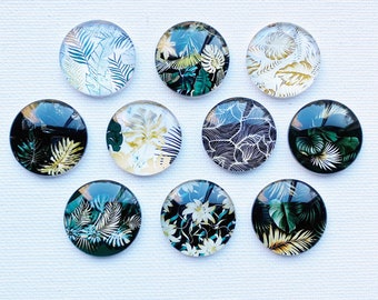 May include: A collection of nine circular glass cabochons featuring various botanical prints. The designs include tropical palm leaves, ferns, and floral patterns in shades of deep green, gold, white, and black, arranged in a grid-like formation.