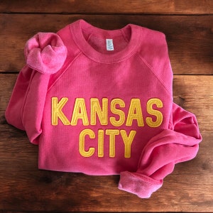 May include: A pink sweatshirt with the words "KANSAS CITY" in yellow embroidered letters.