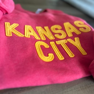 May include: A pink sweatshirt with the words "KANSAS CITY" in yellow embroidered letters.