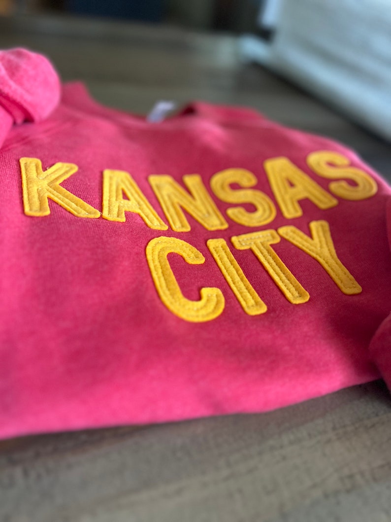 May include: A pink sweatshirt with the words "KANSAS CITY" in yellow embroidered letters.