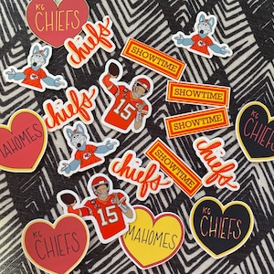 May include: A collection of 12 stickers featuring the Kansas City Chiefs logo, the team mascot, and the names "Chiefs", "Mahomes", and "Showtime". The stickers are in various shapes, including hearts, circles, and rectangles. The stickers are on a black and white patterned surface.