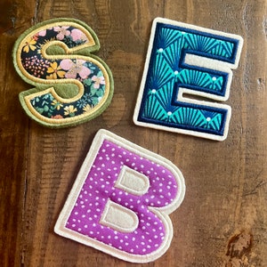May include: Three felt fabric letters, "S", "E", and "B", with different patterns. The "S" is green with a floral pattern, the "E" is blue with a geometric pattern, and the "B" is purple with white polka dots.