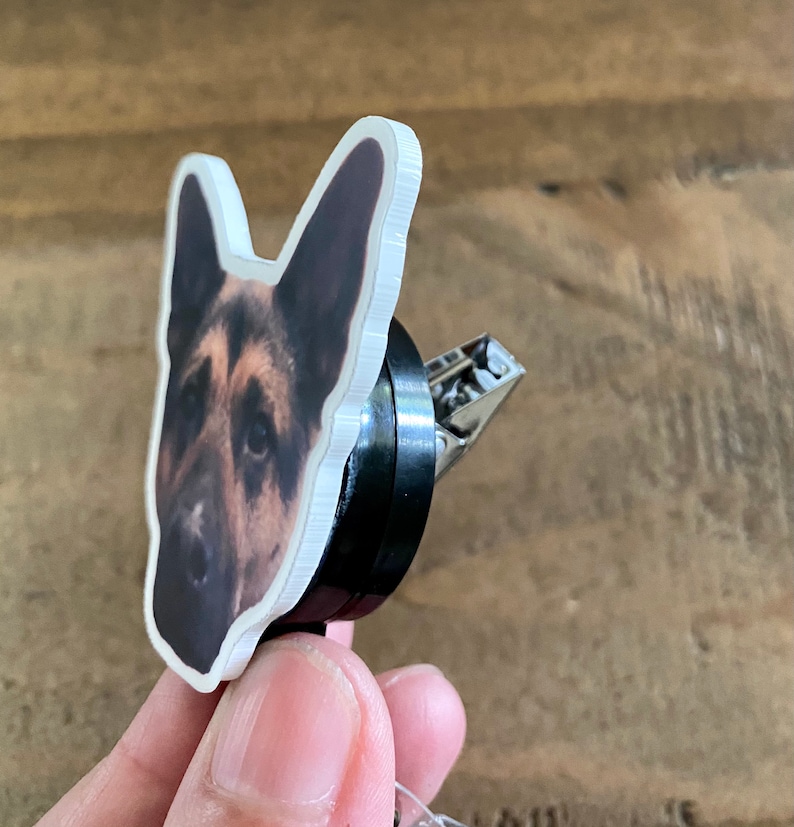 May include: A white plastic badge holder with a printed image of a German Shepherd dog's head. The holder is attached to a black retractable badge reel with a silver clip.