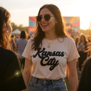 K&ouml;nnte beinhalten: Ein cremefarbenes T-Shirt mit dem Schriftzug "Kansas City" in schwarzer Schrift. Das T-Shirt wird von einer Person getragen, die eine Sonnenbrille und eine Halskette tr&auml;gt. Der Hintergrund zeigt eine Menschenmenge und eine farbenfrohe Struktur.