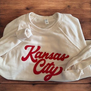 May include: Cream-colored sweatshirt with the words "Kansas City" in red script across the front. The sweatshirt has long sleeves and a crew neck. The garment is folded on a wooden surface.