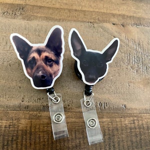 May include: Two black retractable badge holders with a dog's face printed on each. The dog on the left is a German Shepherd with brown and black fur. The dog on the right is black. Both dogs are looking at the camera.