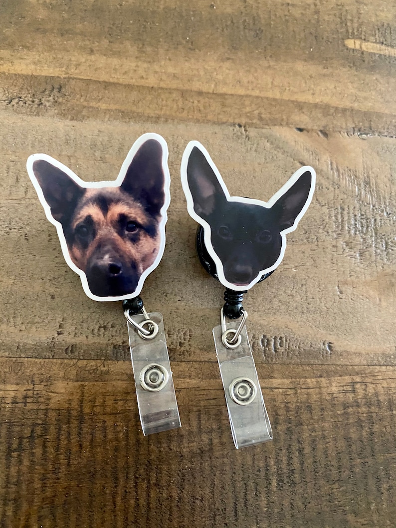 May include: Two black retractable badge holders with a dog's face printed on each. The dog on the left is a German Shepherd with brown and black fur. The dog on the right is black. Both dogs are looking at the camera.