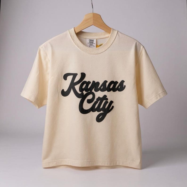 K&ouml;nnte beinhalten: Cremefarbenes T-Shirt mit dem Schriftzug "Kansas City" in schwarzer Schrift. Das Shirt hat kurze &Auml;rmel und einen Rundhalsausschnitt. Das Shirt h&auml;ngt an einem Holzb&uuml;gel.