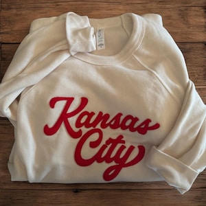 May include: Cream-colored sweatshirt with the words "Kansas City" in red script across the front. The sweatshirt is folded, with the sleeves cuffed. The background is a wooden surface.