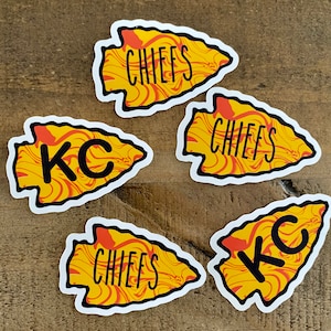May include: Five arrowhead-shaped stickers with a yellow and orange marbled design. The stickers have black outlines and feature the text "KC" and "CHIEFS" in black.