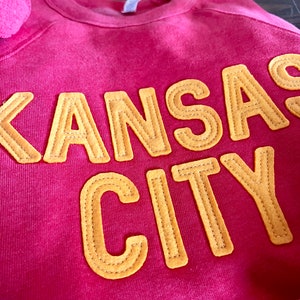 May include: A pink sweatshirt with the words "Kansas City" in yellow felt letters.