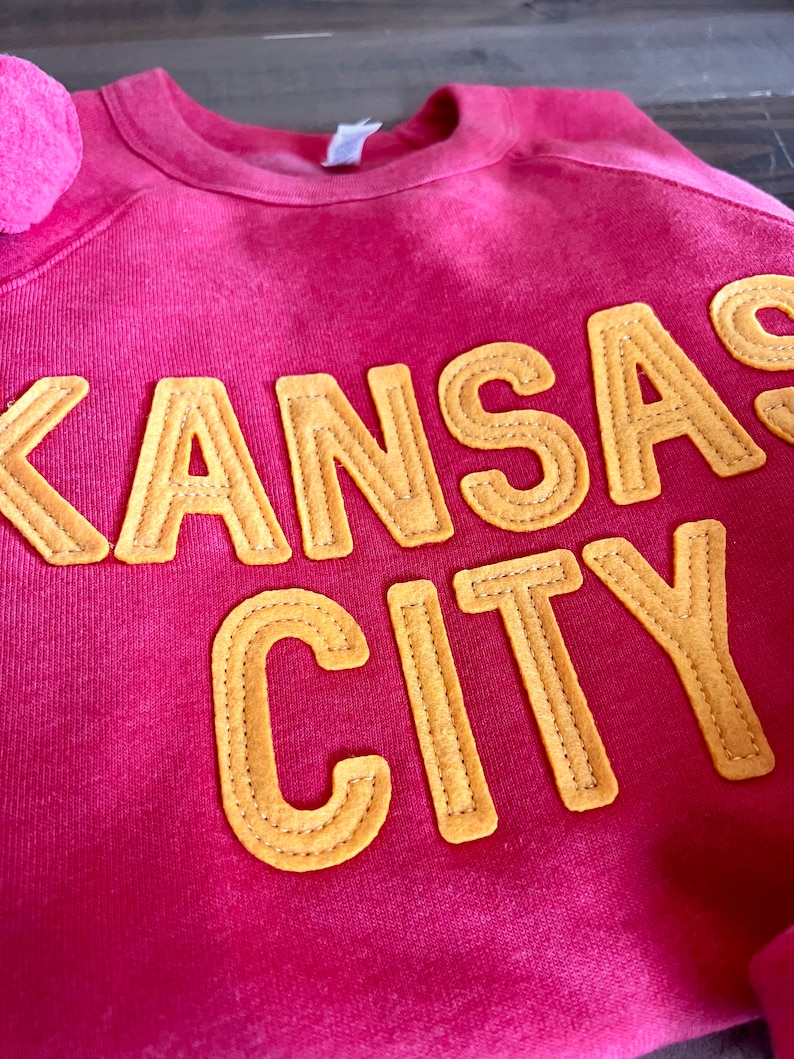 May include: A pink sweatshirt with the words "Kansas City" in yellow felt letters.