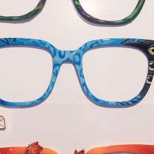 May include: A close-up of a collection of eyeglasses with various frame designs. The central pair features a blue and black pattern with a charm. Other frames include purple, green, and orange designs. The eyeglasses are displayed on a white background.