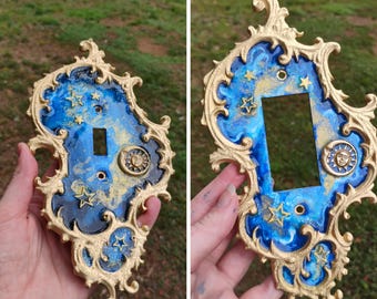 May include: An ornate light switch plate featuring a gold-colored, scrollwork frame surrounding a blue and gold marbled resin center. The design includes embedded gold star shapes and a circular sun-and-moon emblem on the right side.