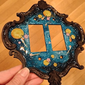 May include: A double light switch plate featuring a dark blue, swirling resin pond design with embedded lily pads, small pink flowers, and koi fish. The plate is encased in an ornate, dark brown, glittery frame with decorative scrollwork edges.