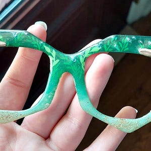 May include: Green-tinted eyeglasses with a nature-inspired design. The frames feature embedded greenery and small, tan mushroom accents. The lower frame edges have a glittery, sand-like texture, creating a unique, artistic look.