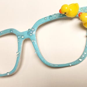 May include: Light blue plastic glasses with small white beads around the frame and two yellow rubber duckies attached to the top.