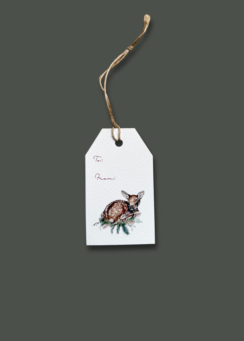 May include: White gift tag with a watercolor illustration of a fawn resting on pine needles. The tag has the words "To:" and "From:" in a handwritten font, and is attached to a natural twine string.