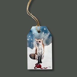 May include: A watercolor gift tag featuring a fox sitting in the snow under a starry sky. The fox has brown and white fur and is perched above a red ornament. The tag is attached to a natural twine string.