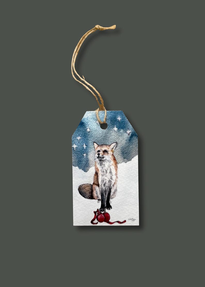 May include: A watercolor gift tag featuring a fox sitting in the snow under a starry sky. The fox has brown and white fur and is perched above a red ornament. The tag is attached to a natural twine string.