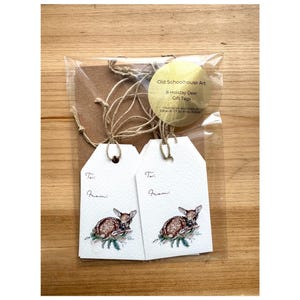 May include: A package of eight holiday deer gift tags. Each white tag features a watercolor illustration of a fawn resting on a branch. The tags are packaged with twine and a brown backing. The package includes a round label with the text "Old Schoolhouse Art".