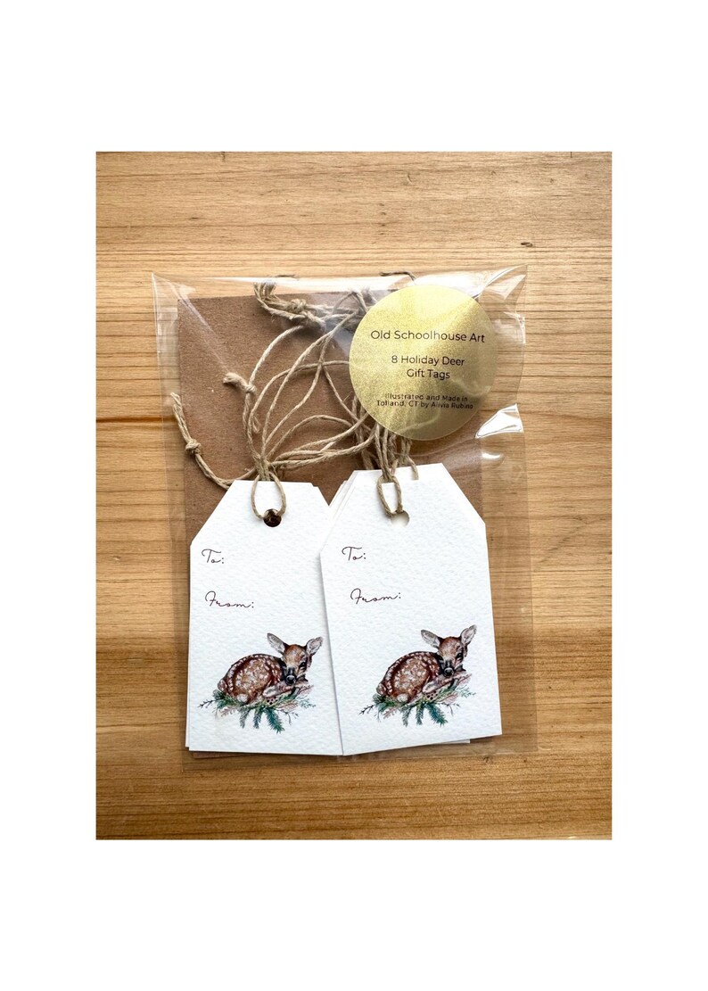 May include: A package of eight holiday deer gift tags. Each white tag features a watercolor illustration of a fawn resting on a branch. The tags are packaged with twine and a brown backing. The package includes a round label with the text "Old Schoolhouse Art".
