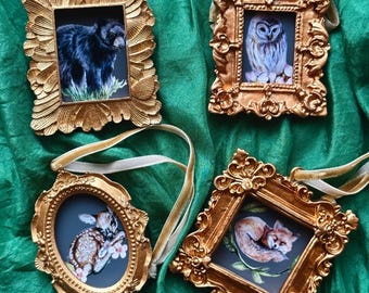 May include: Four decorative hanging ornaments featuring ornate gold-colored frames. The images inside depict a black bear, an owl, a spotted fawn, and a sleeping fox. Each frame includes a cream-colored ribbon loop for hanging purposes.