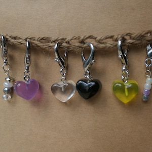 May include: A collection of six heart-shaped charms in various colors, including purple, clear, black, and yellow, suspended from a natural twine. Each charm is attached to a silver-colored clasp, with some featuring beaded accents.