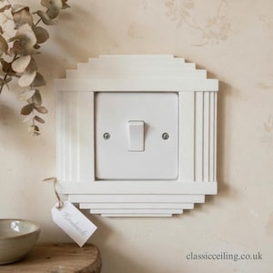 May include: A white, decorative light switch cover with a stepped design and vertical accents. The switch plate has a single toggle switch. A small tag that says "Handmade" is attached. The background is a light beige wall.