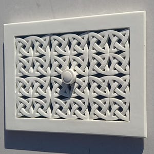 May include: A white rectangular panel featuring a Celtic knot design. The panel has a raised, intricate pattern and a small, round knob in the centre. The frame is also white.