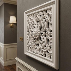 May include: A white, square wall decor piece with an ornate floral and leaf design. The decorative panel is framed and mounted on a gray wall. The intricate relief carving adds a touch of elegance to the interior.