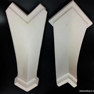 May include: Two white architectural mouldings. One has a V-shaped top, the other a peaked top. Both pieces have a tapered design and a decorative base. The background is black.
