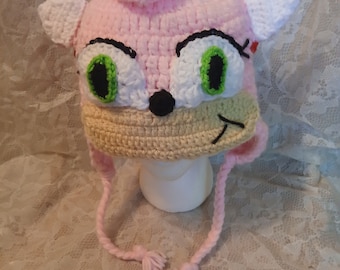 May include: Pink and white crocheted hat with a cartoon face, green eyes, and a black nose. The hat has a long pink tassel.