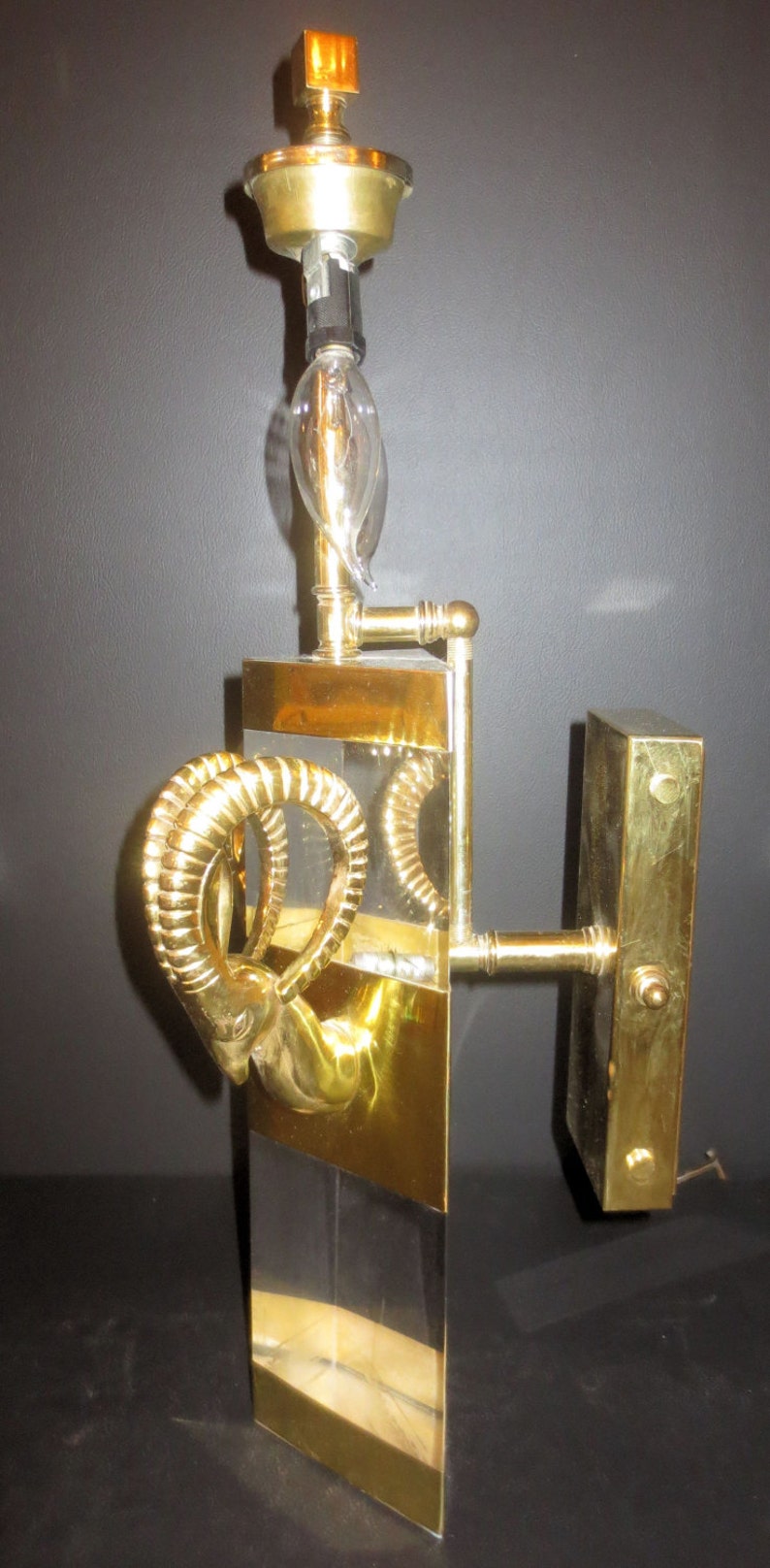 May include: A vintage wall sconce with a brass finish, featuring a ram's head design. The sconce has a clear glass section and a light bulb. The fixture is approximately 18 inches tall.