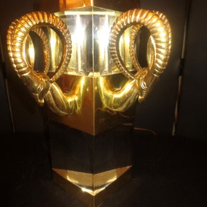 May include: A gold-colored table lamp with a geometric design. The lamp features two ram's head sculptures on the sides. The lamp has a rectangular base and a gold and black color scheme.